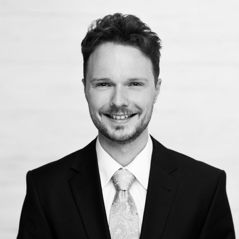 Mann in Anzug mit Krawatte, lächelt vor einem hellen, neutralen Hintergrund.